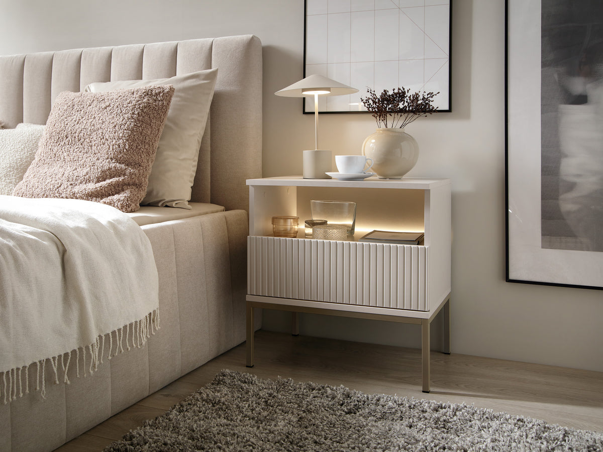White bedside cabinet with vertical ribbed drawer and open shelf beside bed
